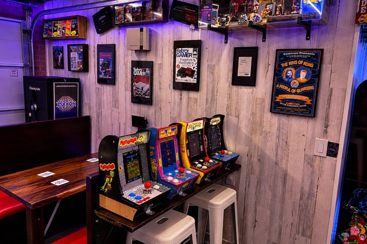 The booth's red seat and dark wood table at the left, beside a counter with four mini arcade cabinets and two white stools, under shelves of boxed games, mounted vintage consoles and framed posters on the white wall.
