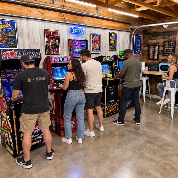 Friends playing the Midway, NBA Jam and Street Fighter II cabinets under the neon ARCADE sign and framed posters, with a friend on a white stool at the counter.