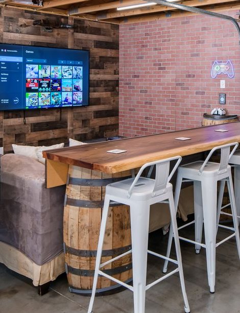 A long live-edge wooden table at bar height on an oak barrel, with white metal stools, set right behind the sectional and facing a television on the wall, with a small neon controller sign beside it.