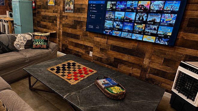 The end of the grey sectional and the stone-topped coffee table with a checkers board set out, facing the large television on the wall.