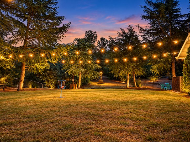 A Boulder Oak disc golf basket on the lawn at dusk, under strings of lights between the pines, with a picnic table and turquoise chairs at the edge of the grass.