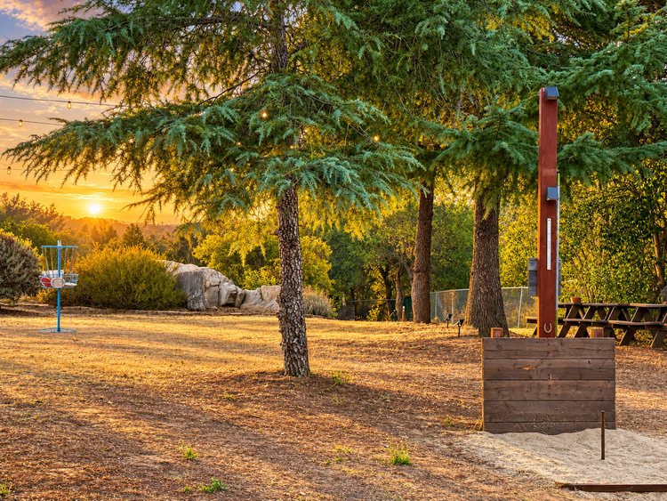 The far pit at golden hour: a sand pit and stake in front of a wooden backboard beside a tall post under the cedars, with the picnic tables and a Boulder Oak disc golf basket beyond.