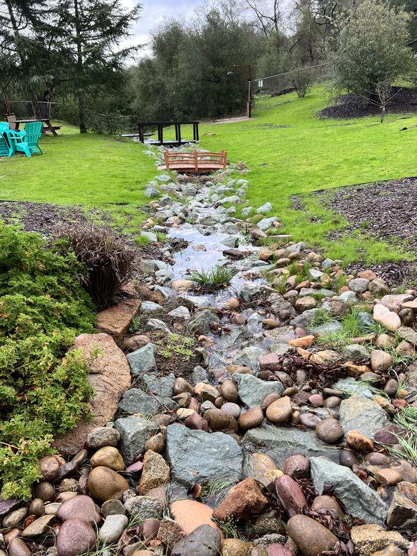 Water running down a rocky stream bed between green banks, with turquoise Adirondack chairs, the small arched garden bridge and the Fairytale Bridge in the distance.