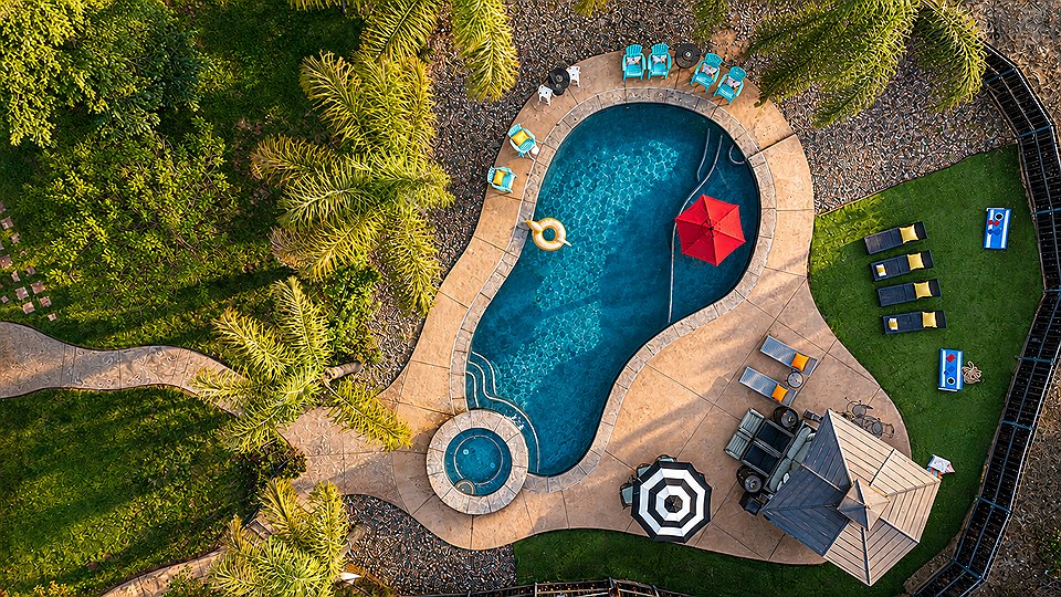 The Oasis from above: the curved pool with the round spa at one end, a red umbrella and a striped one, loungers on the deck and palms around the edge.