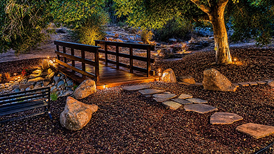 The Fairytale Bridge over the creek bed, lit up at dusk, with flagstone steps, a bench and a lit oak beside it.