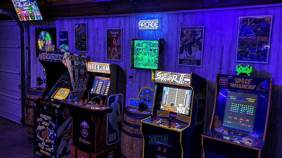 Four arcade cabinets along the wall under blacklight: Midway, NBA Jam, Street Fighter II and Space Invaders, with oak barrels between them, a lit dart board on the left and the neon ARCADE sign above framed posters.
