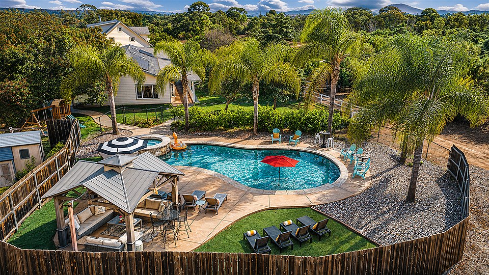 The whole Oasis from the air: the pool and spa, the shaded pavilion, lawn and loungers, all inside the fence with palms along it.