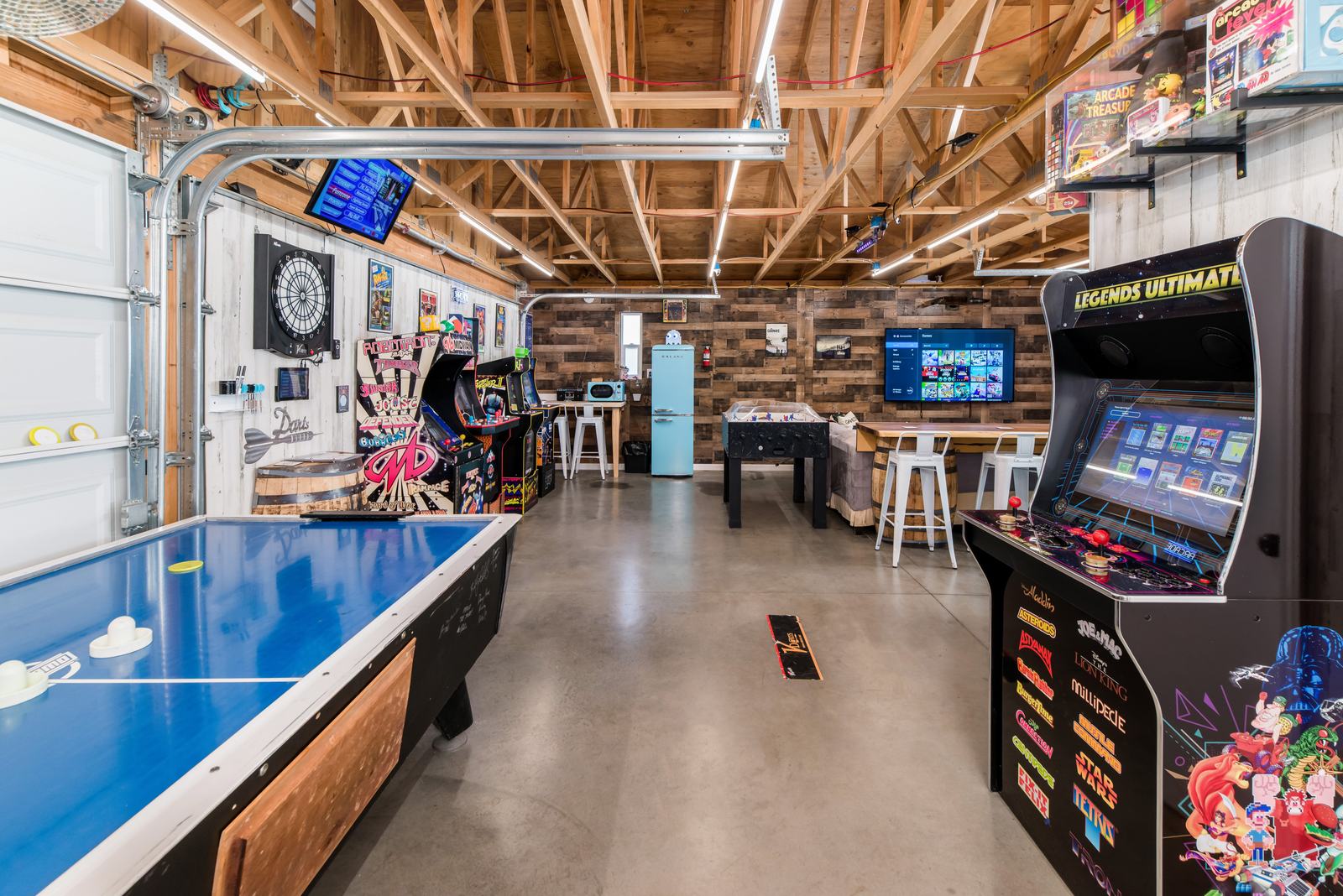 The pub-style arcade: an air hockey table, arcade cabinets, foosball, a dartboard and screens under an open timber roof.