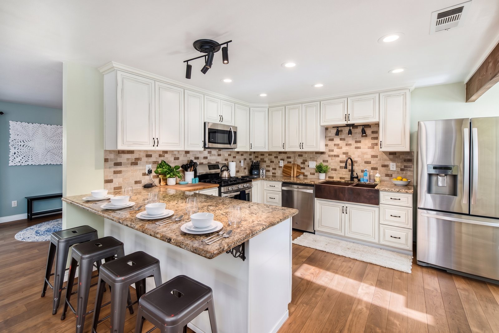 The bright white kitchen with a breakfast bar and stools, stainless appliances and a stone backsplash.