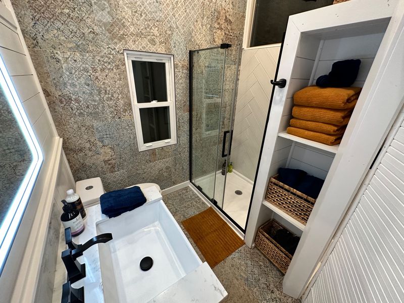 The bathroom: a white vanity beside a lit mirror, a glass shower with black fittings, patterned tile walls and open shelves stacked with mustard and navy towels.