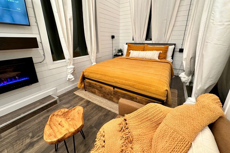 A queen bed in mustard and white bedding between tall curtained windows, with an electric fireplace glowing on the wall and a live-edge side table in front.