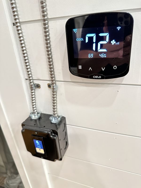 A smart thermostat reading 72 degrees on a white shiplap wall, with silver metal conduit running down to a black switch box.