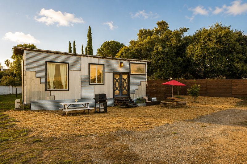The tiny home at golden hour: corrugated steel siding in mixed panels, black-framed windows, black double doors with leaded glass under a lantern, and a picnic table and red umbrella in front.