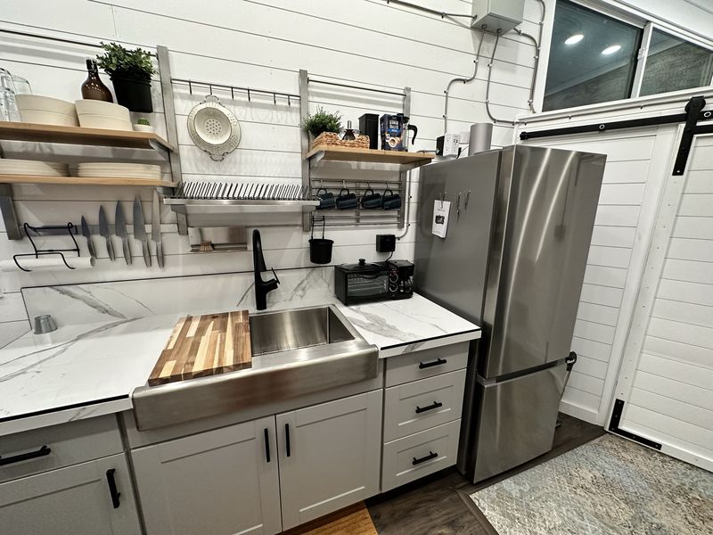 The kitchenette: a stainless farmhouse sink with a butcher-block board across it, white marble-look counters, open shelves of plates and mugs, knives on a magnetic rail, a toaster oven and a stainless fridge.