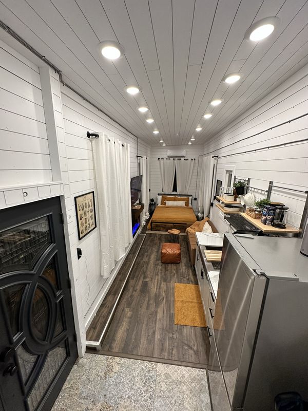 Looking down the full length of the tiny home from the front doors, past the fridge and kitchenette to the lounge and the bed at the far end, under a row of recessed lights.