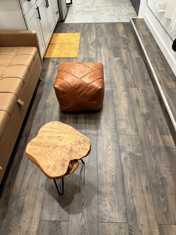 A tan sofa along one wall, a leather pouf and a live-edge wooden side table on hairpin legs, on a wood floor.