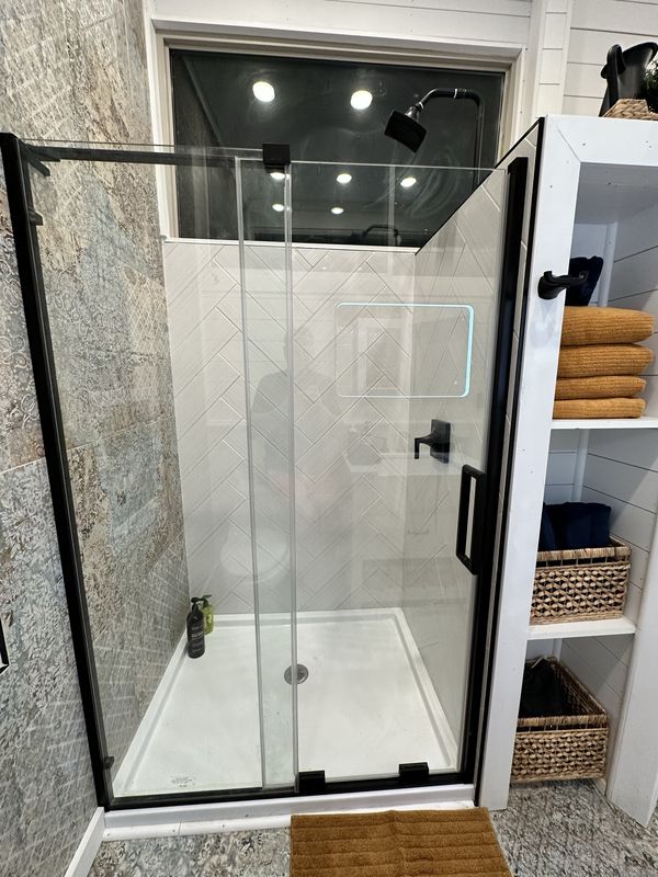 A full-size glass shower with black fittings and white herringbone tile, beside shelves of towels and baskets.