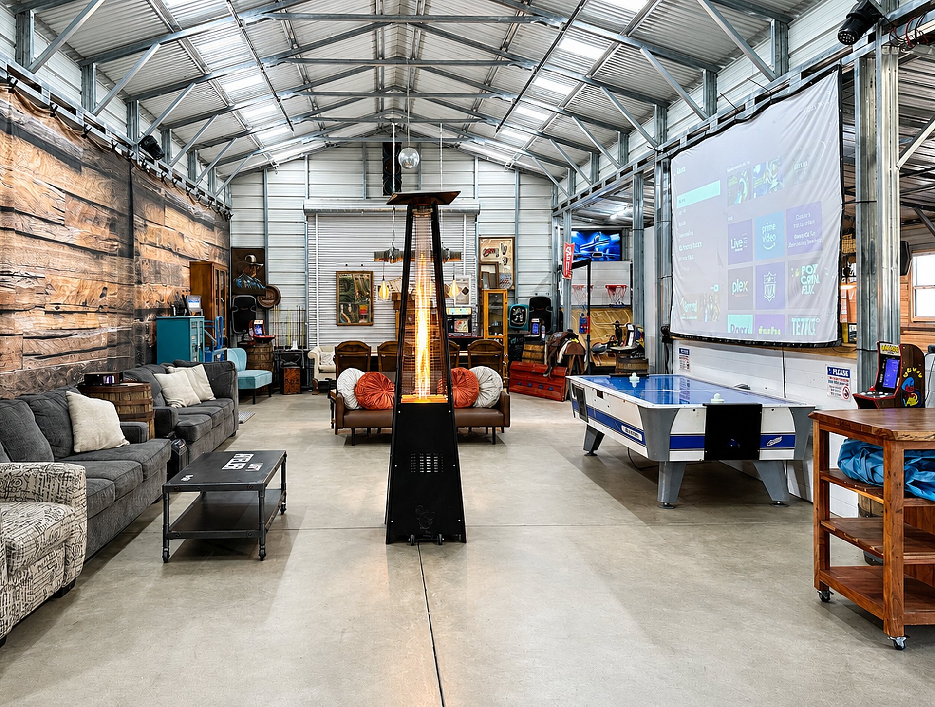 The barn from end to end under the steel trusses: grey sectionals, a lit patio heater, air hockey, the projection screen and arcade games along the walls.