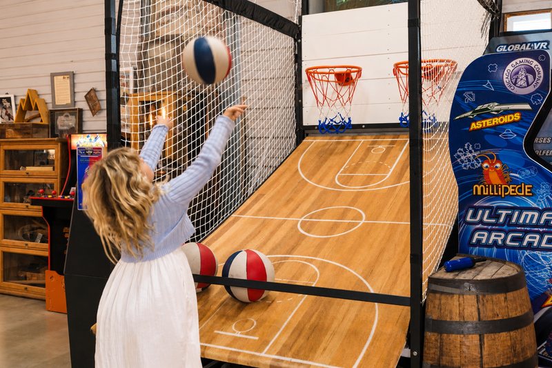 A girl shooting at the two hoops of Legend&rsquo;s Stadium Basketball, with the Ultimate Arcade cabinet beside it.