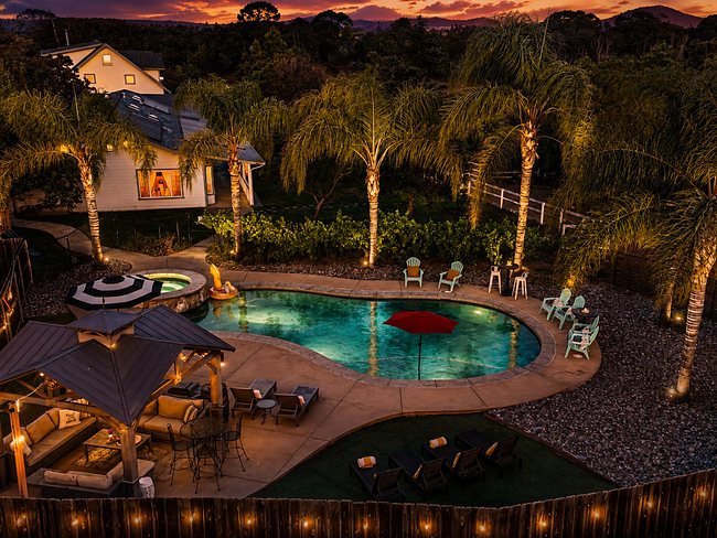 The Oasis after dark: the pool and spa lit from below, string lights along the fence and the palms uplit, the pavilion glowing beside the water.