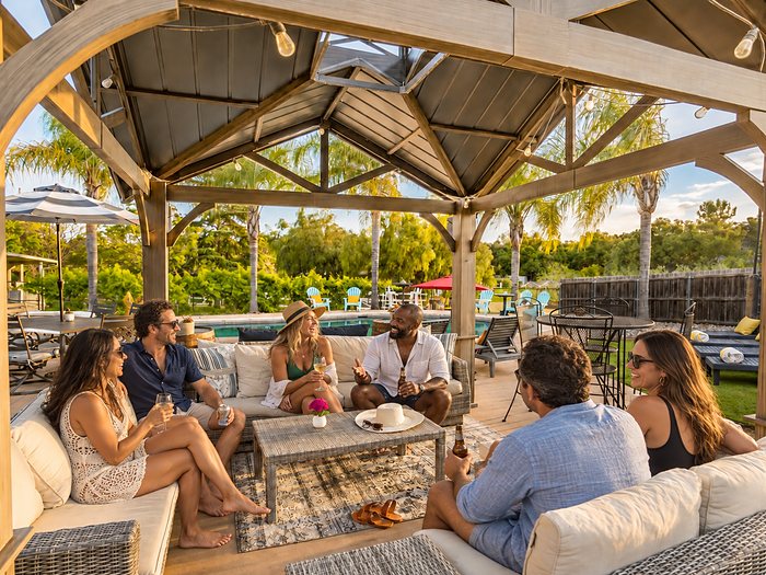 Friends talking on the cushioned sofas under the wooden pavilion beside the pool.